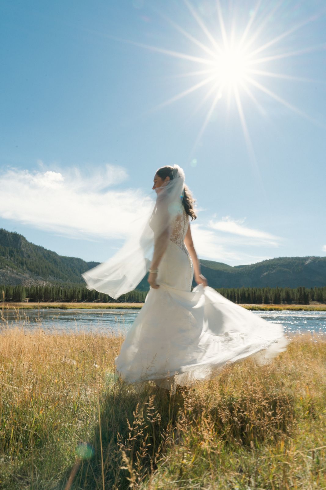 A bride in tall grass beside a mountain lake, veil blowing, sun overhead